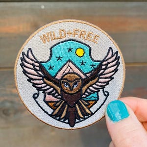 May include: A beige embroidered patch with a brown owl flying in front of a turquoise mountain scene. The text "WILD+FREE" is embroidered in brown around the top of the patch.