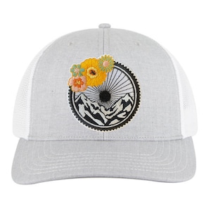 May include: A grey and white trucker hat with an embroidered patch of a mountain scene with a sun and flowers. The patch is black, white, and yellow.