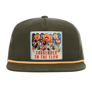 May include: An olive green baseball cap with a flat brim and a yellow cord trim. A rectangular patch on the front features an embroidered floral design in orange, blue, and white, with the text "SURRENDER TO THE FLOW" in red lettering.
