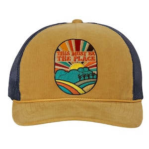May include: A mustard yellow corduroy trucker hat with a black mesh back. The front of the hat features an embroidered patch with a sunset scene and the text "This must be the place".