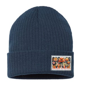 May include: Navy blue knit beanie with a rectangular patch featuring a floral design in orange, red, and yellow. The patch has the text "SURRENDER TO THE FLOW" in white. The beanie is ribbed.