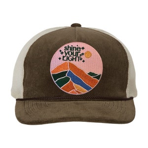 May include: A brown corduroy trucker hat with a white mesh back. The hat features a circular patch with a mountain landscape design and the text "shine your light". The patch includes pink, orange, blue, and green colours.