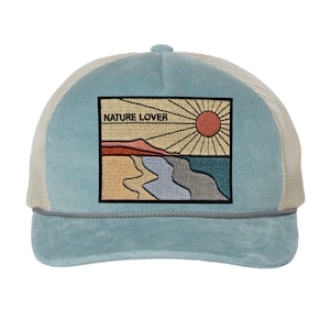 May include: A light blue and white trucker hat with a corduroy front and a mesh back. The hat features an embroidered patch with a sunset scene and the text "NATURE LOVER".