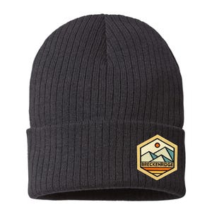 May include: A charcoal grey ribbed knit beanie featuring a hexagonal patch on the cuff. The patch displays a mountain landscape graphic with a sun, blue peaks, and orange stripes, with the text BRECKENRIDGE printed in black capital letters.