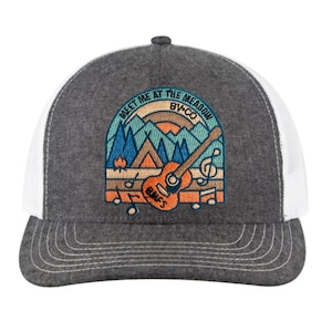 Può includere: Un cappello da camionista grigio e bianco con un ricamo che rappresenta una scena di montagna con un falò, una tenda, una chitarra e il testo "Meet Me At The Meadow BWCO".