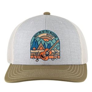 May include: A grey trucker cap with a white mesh back and a brown peak. The front has an embroidered patch depicting a guitar, mountains, and the text "MEET ME AT THE MEADOW BV-CO". Musical notes and a campfire are also shown.