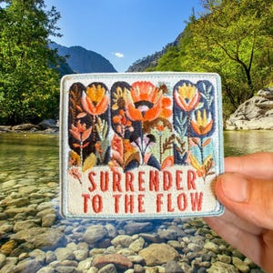May include: A rectangular embroidered patch with a light cream background featuring a colorful floral design in shades of orange, yellow, and dark blue. The text "SURRENDER TO THE FLOW" is stitched in orange letters at the bottom of the patch.