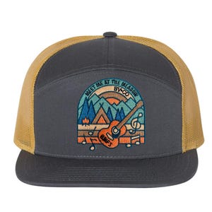 May include: A grey and gold trucker hat with a patch that says "Meet Me At The Meadow By+Co" and features a mountain scene with a campfire, a tent, and a guitar.