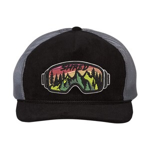 May include: Black and grey trucker hat with a black corduroy front and a grey mesh back. The hat features an embroidered patch of a pair of ski goggles with a mountain scene and the word "SHRED" in a colourful font.
