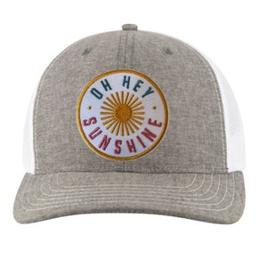 May include: Grey and white trucker cap featuring a circular patch with the text "OH HEY SUNSHINE" in teal, gold, and red. The cap has a curved peak and a mesh back.