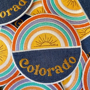 May include: A stack of embroidered patches with a rainbow and a sun design. The patches are navy blue with gold lettering that says "Colorado".