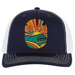 May include: A navy blue and white trucker hat with a retro-style embroidered patch that says "This Must Be The Place". The patch features a colorful sunrise, hills, and trees.