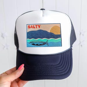 May include: White and navy blue trucker hat with a patch that says "SALTY" and features a stylised fish and a sunset over a mountain range.