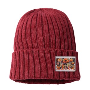 May include: A burgundy ribbed knit beanie with a rectangular patch featuring a floral design and the text "SURRENDER TO THE FLOW". The hat is made of a thick, warm material, perfect for cold weather.