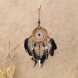 Puede incluir: Un atrapasueños marrón y negro con plumas y cuentas. El atrapasueños está colgado de un cordón marrón.