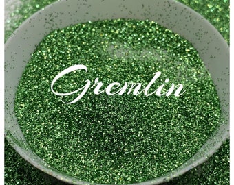 May include: A close-up of a bowl filled with green glitter. The glitter is a fine, sparkly texture and has a slight iridescent sheen. The word "Gremlin" is written in white cursive letters on the glitter.