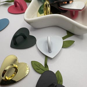 May include: A collection of heart-shaped cabinet pulls in various colors, including gold, black, pink, and blue. Some pulls are metallic, while others have a matte finish. Green fabric leaves are scattered around the pulls.