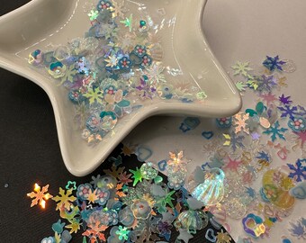 May include: A white star-shaped dish filled with iridescent blue, white, and pink glitter and various shapes, including flowers, stars, hearts, and seashells. The glitter is scattered around the dish on a black surface.
