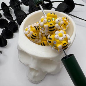 May include: A white skull-shaped bowl filled with yellow and black bee-shaped beads, accented with white flower beads and rhinestones. A green-handled tool is inserted into the beads. Black leaf-shaped decorations are in the background.