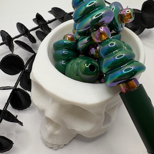 May include: A collection of green and iridescent glass beads in a white skull-shaped container. The beads have a spiral design and are arranged to resemble a decorative item. A dark green pen-like object is also visible.
