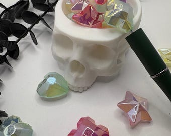May include: A white skull-shaped container filled with iridescent, faceted star and flower-shaped beads. Several loose beads in pink, purple, and green are scattered around the container. A dark green pen is in the background.