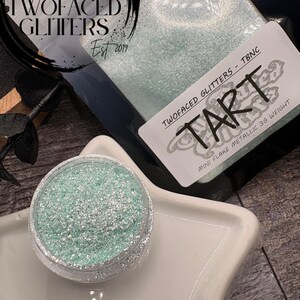 May include: A jar of mint green glitter with silver flakes sits on a white star-shaped dish. The glitter is labeled "TART" and "Mini Flake Metallic 3G Weight" by Two Faced Glitters. A package of glitter is in the background.