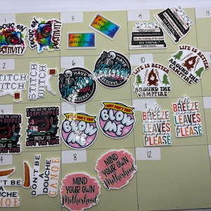 May include: A collection of colorful stickers with various designs and text. Some stickers feature phrases like "Focus on Positivity," "Stitch Witch," and "Life is Better Around the Campfire." Other stickers have humorous sayings and graphics.