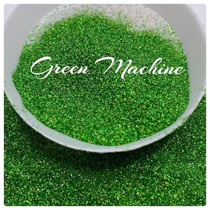 May include: A close-up of a bowl filled with green glitter. The glitter is very fine and has a slight iridescent sheen. The text "Green Machine" is written in white letters across the top of the glitter.