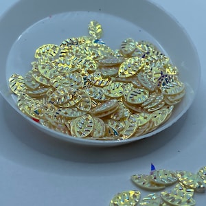 May include: A bowl filled with gold leaf-shaped embellishments. The embellishments have a textured surface and a iridescent finish.