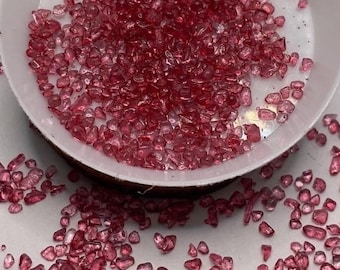 May include: A white bowl filled with small, translucent, ruby-red gemstones. The gemstones are scattered around the bowl on a white surface. The gemstones appear to be tumbled and have irregular shapes. The bowl is made of a white material.