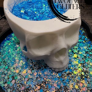 May include: A white skull-shaped container filled with bright blue glitter, resting on a tray of iridescent glitter. The glitter has a mix of shapes and sizes, creating a sparkling effect. The container has the text "TWO FACE GLITTERS" on it.