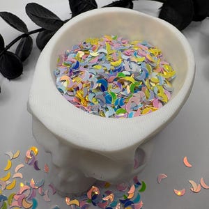 May include: A white skull-shaped bowl filled with iridescent crescent moon-shaped sequins. The sequins have a variety of colors, including yellow, blue, pink, and green. Black decorative leaves are in the background.