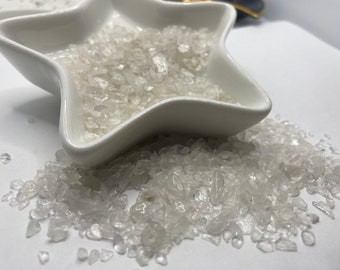 May include: A white, star-shaped ceramic dish filled with small, clear, crushed crystals. Some crystals are scattered around the dish on a white surface. The crystals appear to be quartz or a similar mineral.