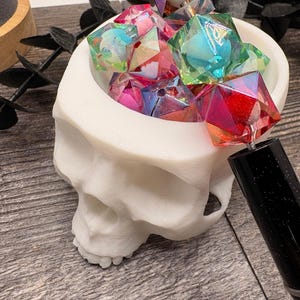 May include: A white skull-shaped container filled with colorful, geometric resin dice. The dice are various colors, including pink, blue, green, and purple, with iridescent and glittery effects. A black pen is in the foreground.