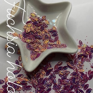 May include: A white star-shaped dish filled with iridescent pink and orange leaf-shaped sequins. The sequins are scattered around the dish on a white surface. The text "$2 - .5 ounce" is written above the dish.