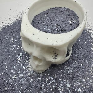 May include: A white skull-shaped bowl filled with dark gray glitter. The glitter spills out onto the surface, creating a textured effect. The bowl has small black dots on the surface.