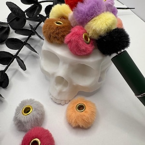 May include: A collection of colorful, fluffy pom-poms with gold eyelets, arranged in a white skull-shaped container. The pom-poms are in various colors, including red, purple, yellow, and pink. A dark green pen is also visible.