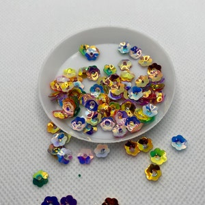 May include: A small white bowl filled with iridescent flower-shaped sequins in shades of yellow, orange, blue, and green. The sequins are scattered around the bowl on a white surface.