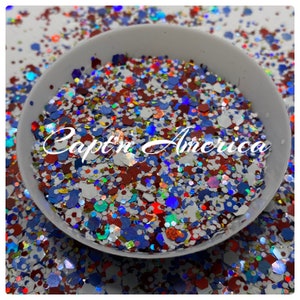 May include: A white bowl filled with red, white, and blue glitter. The glitter is a mix of different sizes and shapes, and it has a holographic finish. The text "Captain America" is written in black letters on the glitter.