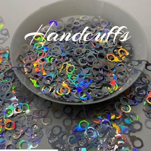 May include: A bowl filled with iridescent holographic glitter shaped like handcuffs. The glitter is scattered around the bowl on a white surface.