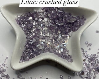 May include: A white star-shaped dish filled with lilac crushed glass. The dish is surrounded by more lilac crushed glass on a white surface. The text "$2 - 1oz Lilac: crushed glass" is written above the dish.