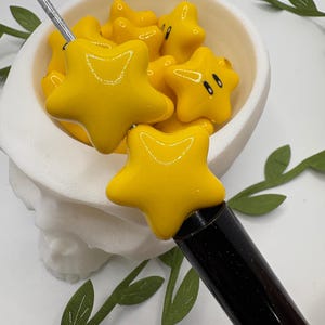 May include: A yellow star-shaped dab tool with a black handle, and a bowl filled with yellow star-shaped objects. The objects have black eyes and a white bowl. The background is white with green leaf accents.