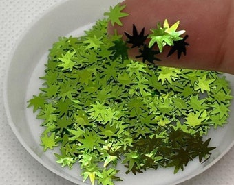 May include: A close-up of a white dish filled with shiny, green leaf-shaped sequins. A finger holds a few of the sequins, some of which are black. The sequins are scattered across the dish, creating a textured, reflective surface.