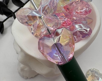 May include: A pen with a faceted, iridescent heart-shaped charm and a green barrel. The pen is surrounded by other heart-shaped and geometric iridescent charms in a white bowl. The charms have pink, purple, and yellow hues.
