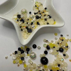 May include: A white bowl filled with a variety of small, round, and flat embellishments in black, white, and yellow. The embellishments are scattered around the bowl and on a white surface.