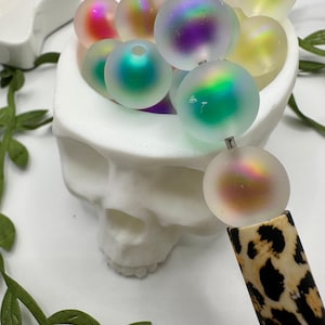 May include: A white skull-shaped container filled with iridescent, round beads. A leopard print pen is in the foreground, with a bead attached. Green leaf garland and a star-shaped dish are also visible.