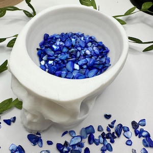 May include: A white ceramic skull-shaped bowl filled with blue iridescent sea shell chips. The bowl is sitting on a white surface with more blue shell chips scattered around it.