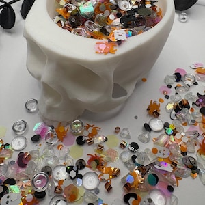 May include: A white skull-shaped bowl filled with Halloween-themed sequins and embellishments. The mix includes orange spiders, ghosts, and iridescent shapes. The bowl sits on a white surface, with black decorative leaves in the background.