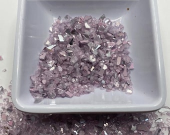 May include: A small white square dish filled with light purple crushed glass. The dish is sitting on a white surface. The text "$2 - 1oz Lt. Purple crush glass" is written above the dish.