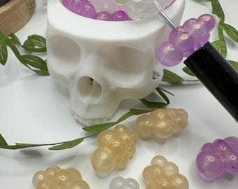 May include: A white skull-shaped container filled with small, iridescent purple, green, and white beads.  The beads are arranged in clusters and are scattered around the container.  There are also several clusters of beads in various colors on a white surface.
